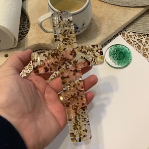 Puede incluir: Un objeto decorativo transparente en forma de cruz con piedras marrones y rosas incrustadas. La cruz se sostiene en una mano, con una taza de té y un rollo de toallas de papel en el fondo. También se ve un pequeño objeto circular verde y blanco.