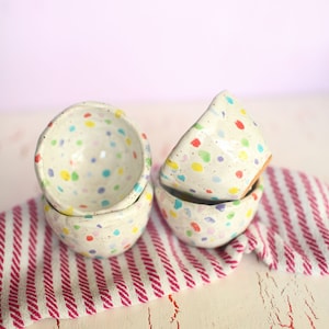 May include: Four small, speckled ceramic bowls are stacked on a red and white striped cloth. The bowls are off-white with colorful polka dots. The bowls are handmade and have a rustic, artisanal feel.
