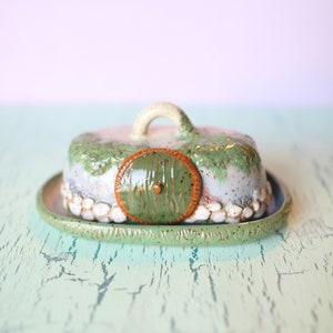 Pu&ograve; includere: Un burriera in ceramica con coperchio, caratterizzata da un design stravagante. Il coperchio &egrave; a forma di casa di hobbit con una porta verde e una maniglia. Il piatto &egrave; dipinto in tonalit&agrave; di verde, blu e bianco, con un aspetto strutturato e rustico.