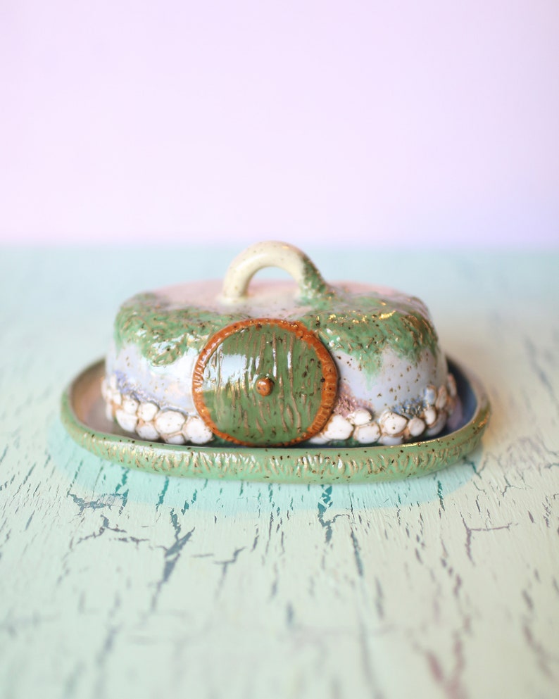 Pu&ograve; includere: Un burriera in ceramica con coperchio, caratterizzata da un design stravagante. Il coperchio &egrave; a forma di casa di hobbit con una porta verde e una maniglia. Il piatto &egrave; dipinto in tonalit&agrave; di verde, blu e bianco, con un aspetto strutturato e rustico.