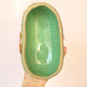 Pu&ograve; includere: Una ciotola in ceramica fatta a mano con un interno turchese macchiato e lucido. Il bordo &egrave; di colore beige chiaro e l'esterno presenta un design strutturato e scanalato. La ciotola &egrave; di forma ovale.