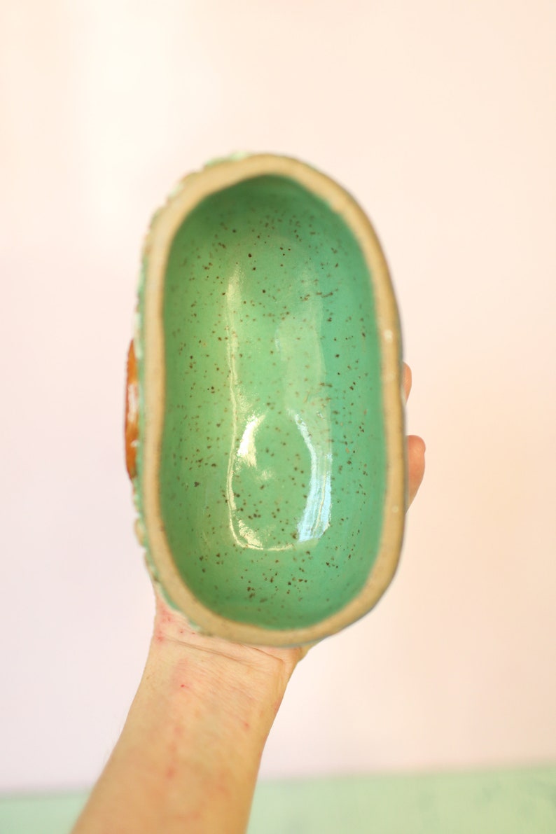 Pu&ograve; includere: Una ciotola in ceramica fatta a mano con un interno turchese macchiato e lucido. Il bordo &egrave; di colore beige chiaro e l'esterno presenta un design strutturato e scanalato. La ciotola &egrave; di forma ovale.