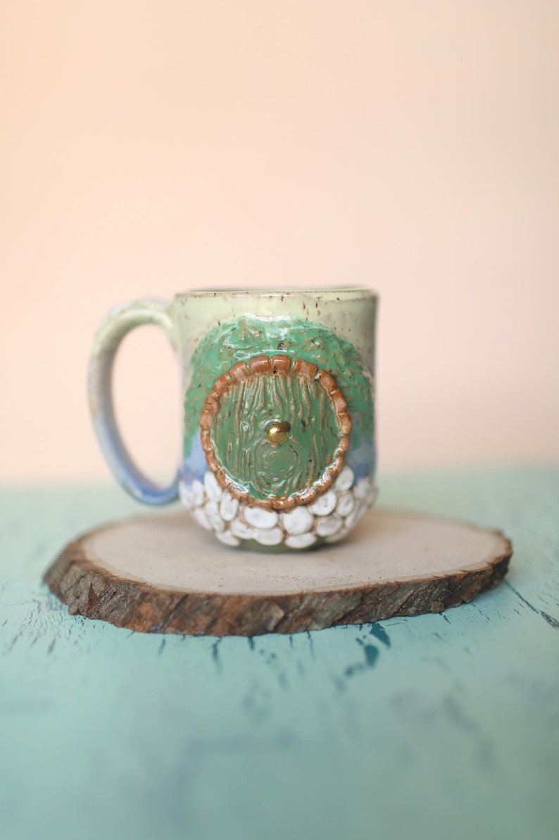 May include: A ceramic mug with a green, brown, and white design. The mug has a round handle and a round, textured design on the front. The design features a small, round, gold-colored knob in the center.