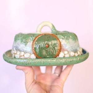 Pu&ograve; includere: Una burriera in ceramica a forma di buco di hobbit. La porta verde &egrave; incorniciata da un bordo marrone. Il coperchio ha una maniglia bianca e dettagli strutturati verdi e bianchi. Il piatto poggia su un piatto rotondo verde.