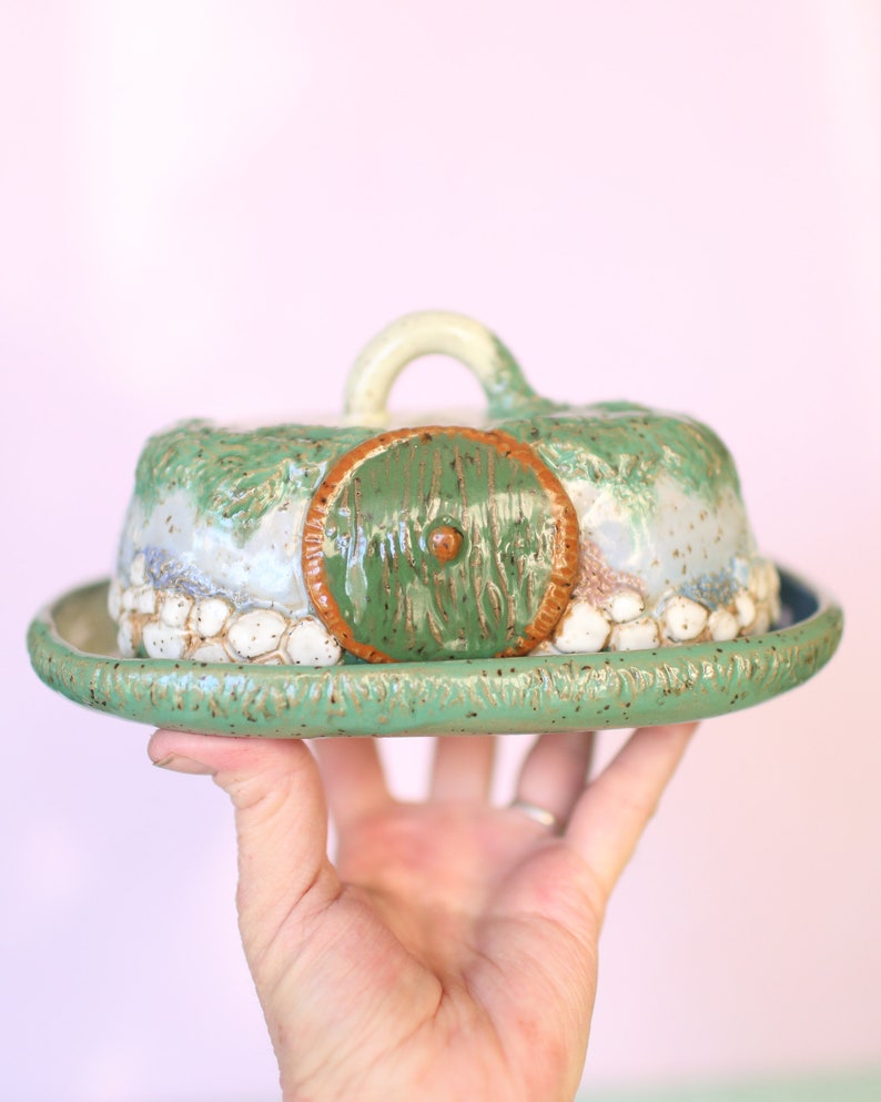 Pu&ograve; includere: Una burriera in ceramica a forma di buco di hobbit. La porta verde &egrave; incorniciata da un bordo marrone. Il coperchio ha una maniglia bianca e dettagli strutturati verdi e bianchi. Il piatto poggia su un piatto rotondo verde.
