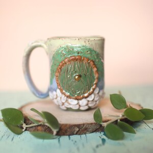 May include: A ceramic mug with a green door design. The door has a brown frame and a gold knob. The mug is decorated with white dots and is sitting on a wooden slice.