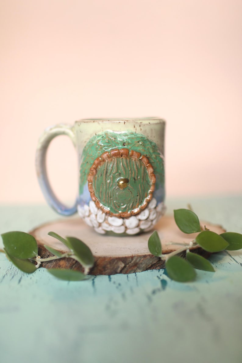 May include: A ceramic mug with a green door design. The door has a brown frame and a gold knob. The mug is decorated with white dots and is sitting on a wooden slice.