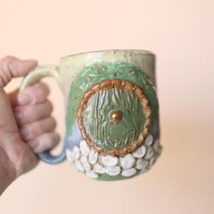 May include: A ceramic mug with a green, brown, and white design. The mug has a round, green door with a gold knob on the front.