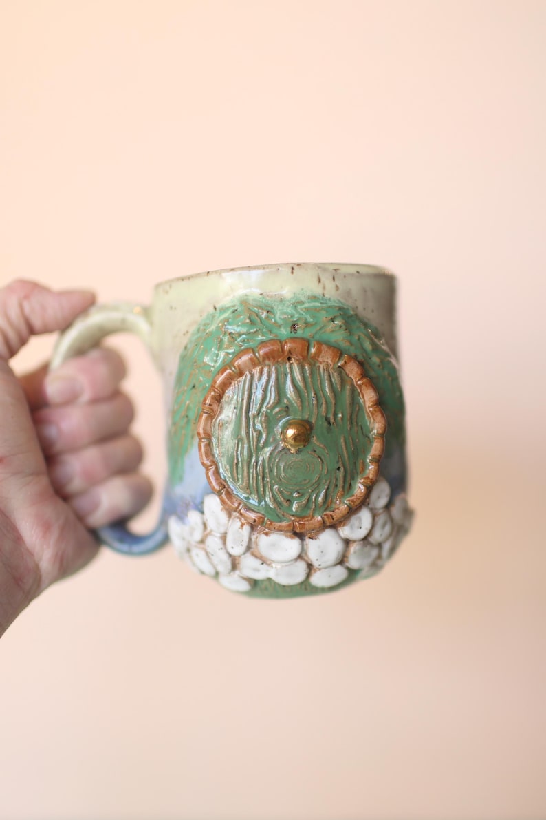 May include: A ceramic mug with a green, brown, and white design. The mug has a round, green door with a gold knob on the front.