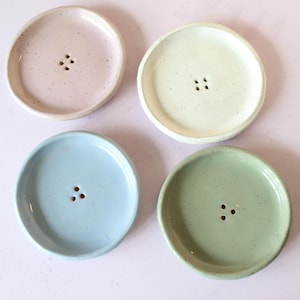 May include: Four ceramic soap dishes in pastel shades: pink, cream, blue, and green. Each dish is round with a raised edge and three drainage holes. The dishes have a speckled texture.