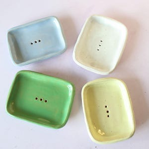 Puede incluir: Cuatro jaboneras de cerámica en colores pastel: azul claro, crema, verde y amarillo. Cada plato es rectangular con esquinas redondeadas y tres agujeros de drenaje. Los platos están dispuestos sobre una superficie clara.