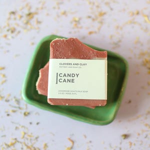 May include: A bar of handmade goat's milk soap with a "Candy Cane" label, resting on a green ceramic dish. The soap is a reddish-brown color. The label includes the brand name "Clovers and Clay" and the soap's weight, 5.5 oz.