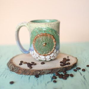 May include: A ceramic mug with a green and brown door design, white speckled glaze, and a gold knob. The mug is sitting on a wooden slice with coffee beans scattered around it.