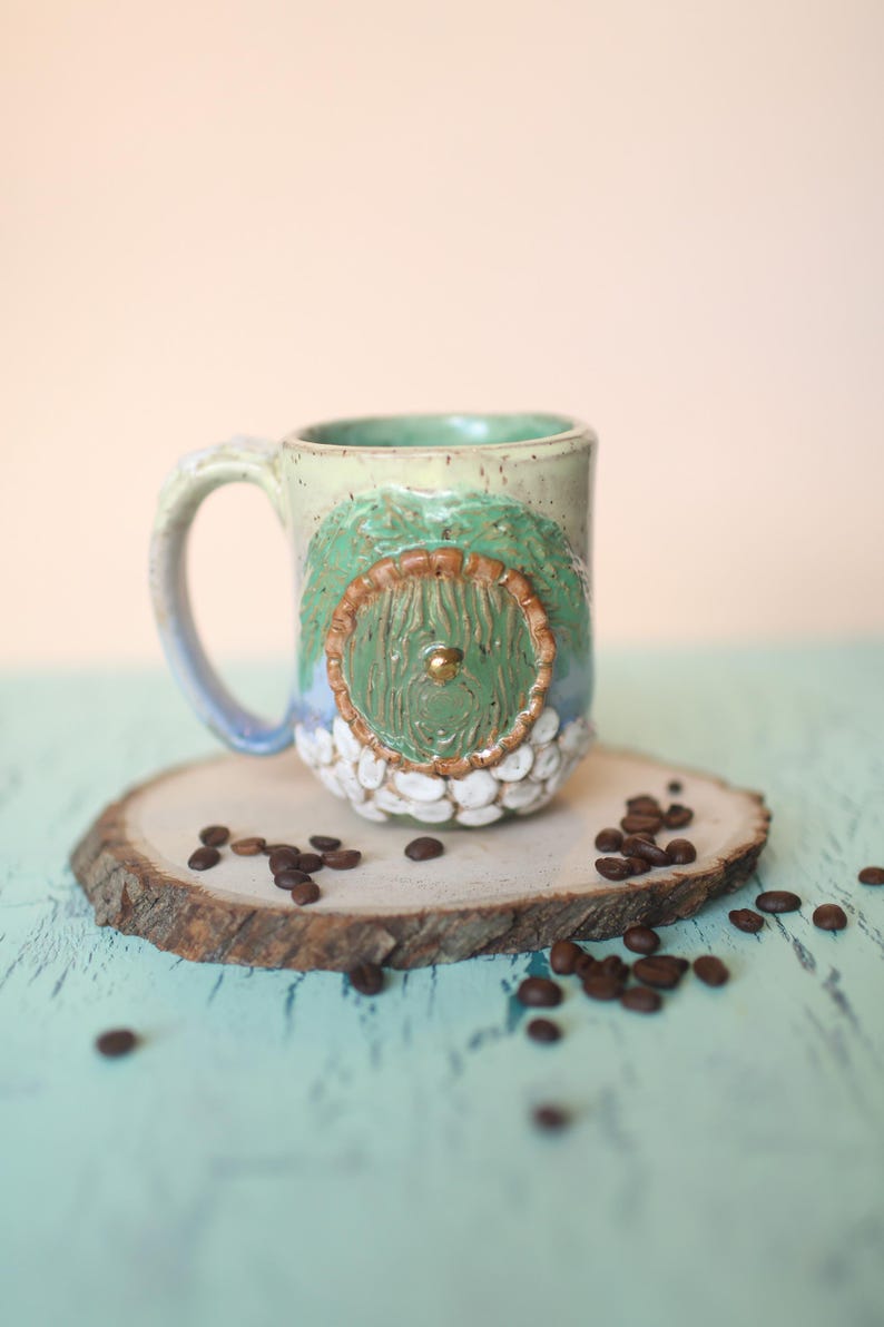 May include: A ceramic mug with a green and brown door design, white speckled glaze, and a gold knob. The mug is sitting on a wooden slice with coffee beans scattered around it.