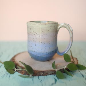 May include: A handmade ceramic mug with a light green, pink, and blue ombre glaze. The mug has a rounded body and a slightly flared lip. The handle is attached to the side of the mug and has a slight curve.