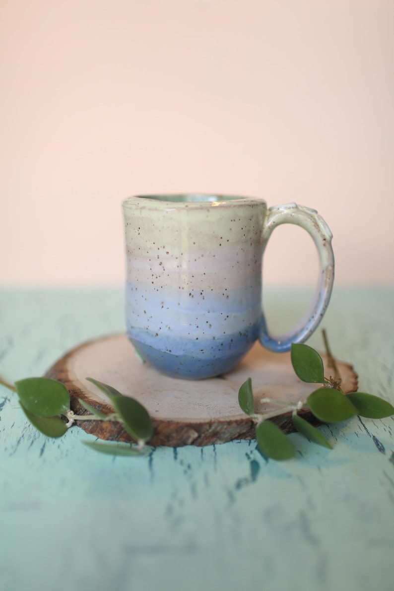 May include: A handmade ceramic mug with a light green, pink, and blue ombre glaze. The mug has a rounded body and a slightly flared lip. The handle is attached to the side of the mug and has a slight curve.