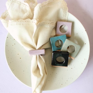 Peut inclure: Une serviette en lin crème avec une bordure en dentelle est maintenue par un anneau de serviette rectangulaire lavande. La serviette repose sur une assiette verte clair mouchetée. Des ronds de serviette carrés supplémentaires en bleu sarcelle, noir et vert sont également présents.