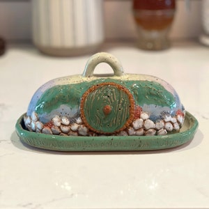 Pu&ograve; includere: Burriera in ceramica fatta a mano con un design stravagante. Il coperchio presenta una porta hobbit verde e marrone, con colline verdi e un cielo azzurro. La base &egrave; di forma ovale verde chiaro.