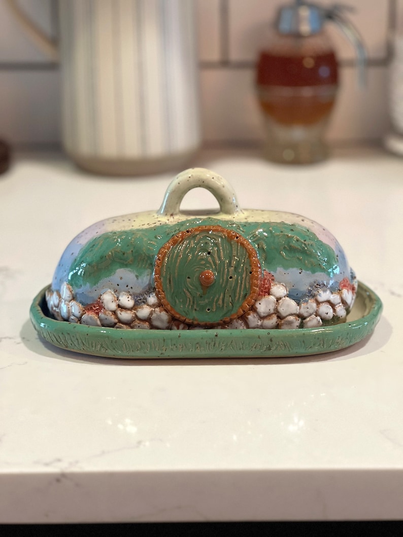 Pu&ograve; includere: Burriera in ceramica fatta a mano con un design stravagante. Il coperchio presenta una porta hobbit verde e marrone, con colline verdi e un cielo azzurro. La base &egrave; di forma ovale verde chiaro.