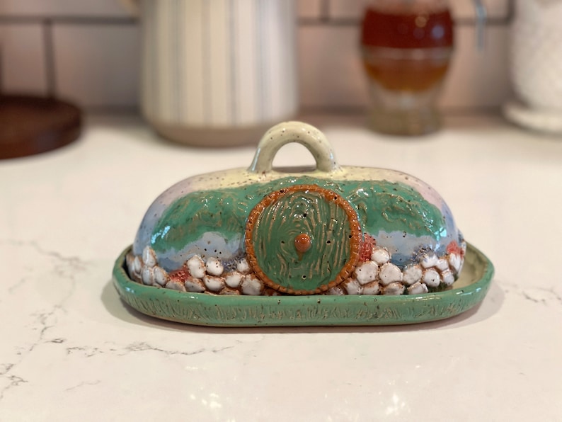 Pu&ograve; includere: Burriera in ceramica fatta a mano con un design stravagante. Il coperchio presenta un paesaggio verde, blu e bianco con una porta rotonda verde. Anche la base e il manico sono verdi. Il piatto &egrave; progettato per contenere il burro.