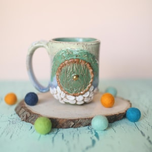 May include: A ceramic mug with a green and brown tree design. The mug has a white glaze with a speckled finish. The tree has a round door with a gold knob. The mug is sitting on a wooden slice with colorful felt balls.