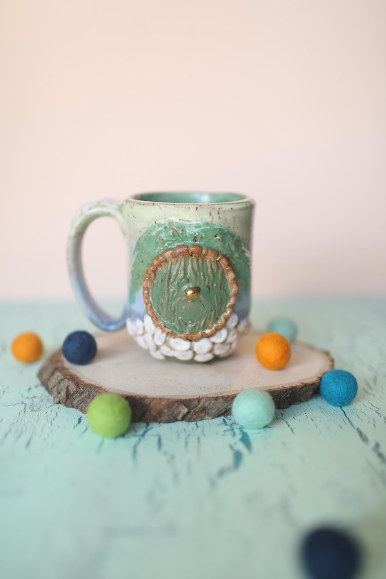 May include: A ceramic mug with a green and brown tree design. The mug has a white glaze with a speckled finish. The tree has a round door with a gold knob. The mug is sitting on a wooden slice with colorful felt balls.