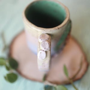 May include: A ceramic mug with a light green interior and a speckled, multi-colored exterior. The mug has a unique handle with three raised bumps.