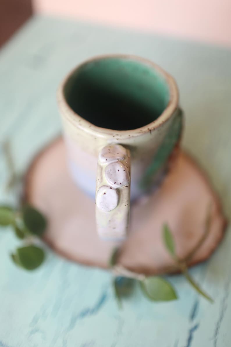 May include: A ceramic mug with a light green interior and a speckled, multi-colored exterior. The mug has a unique handle with three raised bumps.