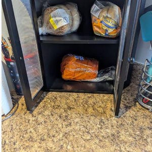 May include: A black cabinet with glass doors, holding packaged bread products. The cabinet has two shelves, with various loaves and rolls visible. The cabinet is open, revealing the contents inside.