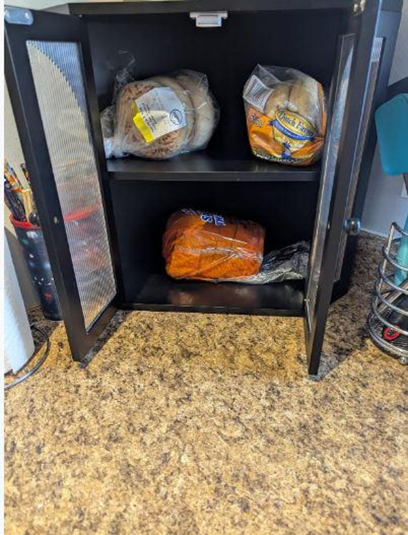 May include: A black cabinet with glass doors, holding packaged bread products. The cabinet has two shelves, with various loaves and rolls visible. The cabinet is open, revealing the contents inside.