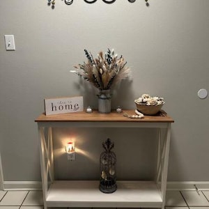 May include: A decorative console table with a light wood top and white base. The table is adorned with a vase of dried flowers, a wooden sign that says "this is home", and a decorative birdcage. A metal wall art piece hangs above the table.