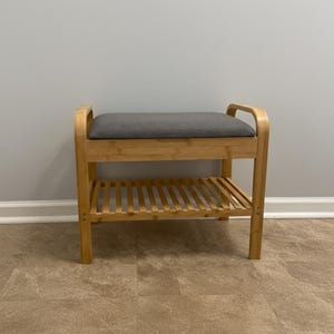May include: A wooden bench with a gray cushioned seat and a slatted shelf below. The bench has a natural wood finish and curved armrests. The bench is designed for seating and storage.