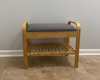 May include: A wooden bench with a gray cushioned seat and a slatted shelf below. The bench has a natural wood finish and curved armrests. The bench is designed for seating and storage.