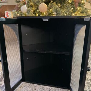 May include: A black cabinet with two doors, each featuring a textured, vertical-lined glass panel. The cabinet is open, revealing a dark interior with a single shelf. The background includes a decorated Christmas tree.