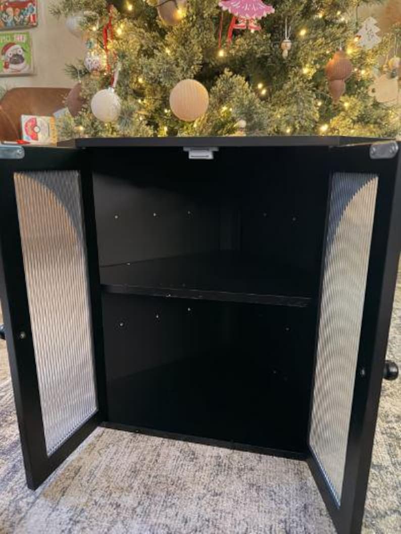 May include: A black cabinet with two doors, each featuring a textured, vertical-lined glass panel. The cabinet is open, revealing a dark interior with a single shelf. The background includes a decorated Christmas tree.