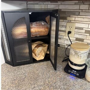 May include: Black cabinet with glass doors, holding loaves of bread. One loaf is in a plastic bag, the other in a paper bag. A scale with a jar and a wooden lid sits on the counter. The cabinet is open, revealing the contents.