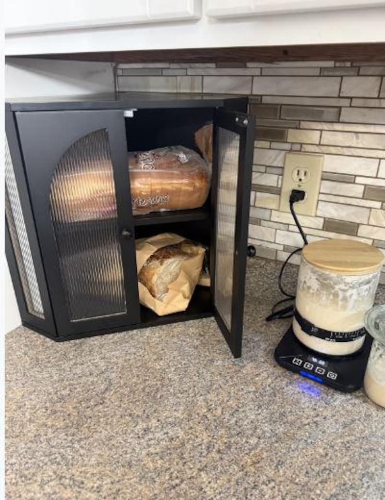 May include: Black cabinet with glass doors, holding loaves of bread. One loaf is in a plastic bag, the other in a paper bag. A scale with a jar and a wooden lid sits on the counter. The cabinet is open, revealing the contents.