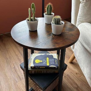 May include: A circular, dark brown side table with a black metal frame. Three small cacti in white pots are on top. The table has a lower shelf with books and a decorative basket.