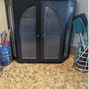 May include: Black corner cabinet with glass-paneled doors. The cabinet has a textured glass design and two round black knobs. A yellow container is on top of the cabinet. A cup of pencils and a utensil holder are on either side.