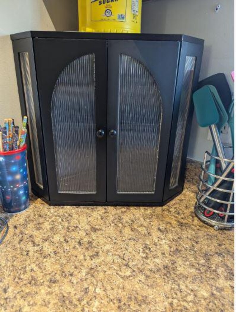 May include: Black corner cabinet with glass-paneled doors. The cabinet has a textured glass design and two round black knobs. A yellow container is on top of the cabinet. A cup of pencils and a utensil holder are on either side.