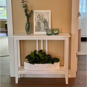 Puede incluir: Una consola blanca con una balda inferior y soportes verticales. La balda superior exhibe un jarrón de cristal verde con vegetación, una imagen enmarcada de jarrones y dos candelabros cuadrados de color verde azulado. Una jardinera blanca con plantas verdes se encuentra en la balda inferior.