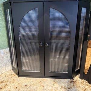 May include: A black corner cabinet with two doors and glass panels. The glass has a vertical ribbed texture. The cabinet has a silver knob on each door. The cabinet is sitting on a speckled countertop.