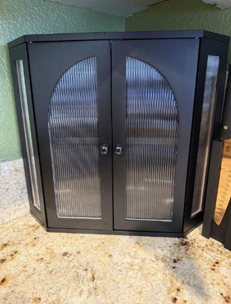 May include: A black corner cabinet with two doors and glass panels. The glass has a vertical ribbed texture. The cabinet has a silver knob on each door. The cabinet is sitting on a speckled countertop.