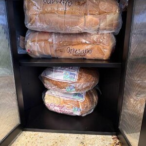 May include: A black cabinet with glass doors, filled with loaves of bread in plastic packaging. The top two shelves hold sliced Artesano bread, while the bottom shelves contain sourdough loaves. The cabinet is open, revealing a countertop.