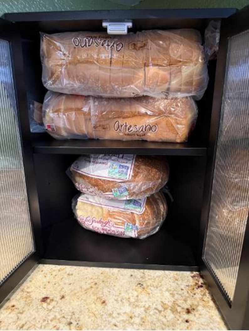 May include: A black cabinet with glass doors, filled with loaves of bread in plastic packaging. The top two shelves hold sliced Artesano bread, while the bottom shelves contain sourdough loaves. The cabinet is open, revealing a countertop.