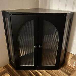 May include: Black corner cabinet with two doors and glass panels. The cabinet has a triangular shape, designed to fit into a corner. The doors have a vertical ribbed pattern, and the cabinet is made of wood.
