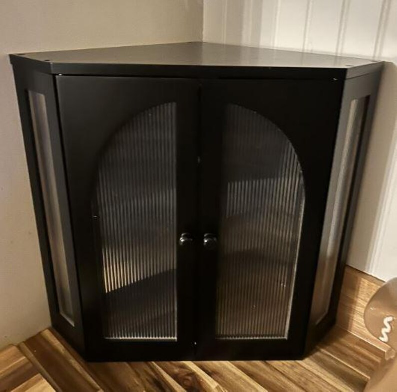 May include: Black corner cabinet with two doors and glass panels. The cabinet has a triangular shape, designed to fit into a corner. The doors have a vertical ribbed pattern, and the cabinet is made of wood.