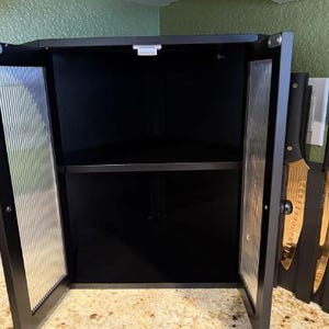 May include: Black corner cabinet with two glass doors, open to reveal a shelf. The glass doors have a vertical ribbed design. The cabinet is mounted under a wooden cabinet and above a speckled countertop. Knives are visible on the right side.