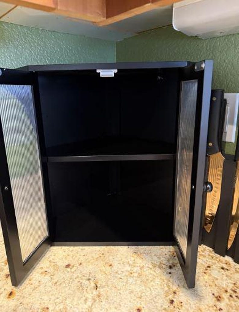May include: Black corner cabinet with two glass doors, open to reveal a shelf. The glass doors have a vertical ribbed design. The cabinet is mounted under a wooden cabinet and above a speckled countertop. Knives are visible on the right side.