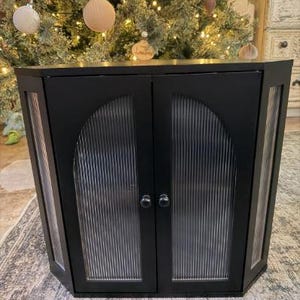 以下が含まれることがあります： 2つのドアとガラスパネルを備えた黒いコーナーキャビネット。キャビネットは、湾曲した上部とシルバーのハードウェアを備えたモダンなデザインです。ガラスパネルは垂直のリブテクスチャです。キャビネットはクリスマスツリーの前に配置されています。